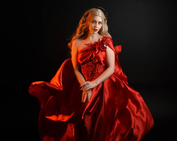 Крупный план портрета красивой блондинки в красном шелковом халате и короне, одетой как древняя мифологическая богиня фантазий. Изящная элегантная поза изолирована на темном фоне студии.