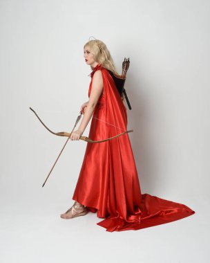 Eski mitolojik fantezi tanrıçası kılığında kırmızı ipek toga elbisesi ve taç giymiş güzel sarışın modelin tam boy portresi. Beyaz stüdyo arka planında izole edilmiş okçuluk yayı ve ok silahları tutan zarif bir poz..