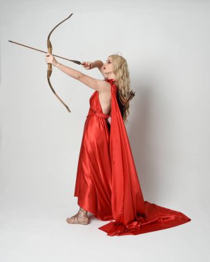 Eski mitolojik fantezi tanrıçası kılığında kırmızı ipek toga elbisesi ve taç giymiş güzel sarışın modelin tam boy portresi. Beyaz stüdyo arka planında izole edilmiş okçuluk yayı ve ok silahları tutan zarif bir poz..