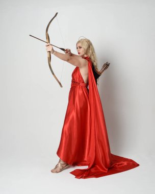 Eski mitolojik fantezi tanrıçası kılığında kırmızı ipek toga elbisesi ve taç giymiş güzel sarışın modelin tam boy portresi. Beyaz stüdyo arka planında izole edilmiş okçuluk yayı ve ok silahları tutan zarif bir poz..