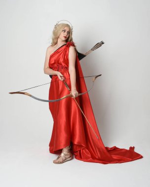 Eski mitolojik fantezi tanrıçası kılığında kırmızı ipek toga elbisesi ve taç giymiş güzel sarışın modelin tam boy portresi. Beyaz stüdyo arka planında izole edilmiş okçuluk yayı ve ok silahları tutan zarif bir poz..