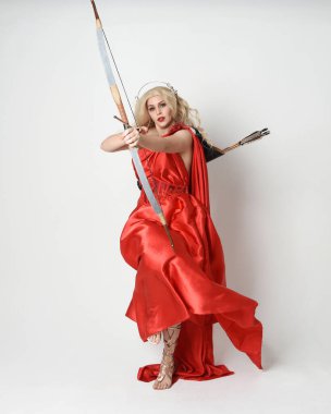 Eski mitolojik fantezi tanrıçası kılığında kırmızı ipek toga elbisesi ve taç giymiş güzel sarışın modelin tam boy portresi. Beyaz stüdyo arka planında izole edilmiş okçuluk yayı ve ok silahları tutan zarif bir poz..