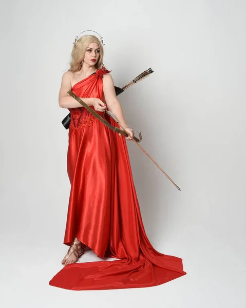 stock image Full length portrait of beautiful blonde model dressed as ancient mythological fantasy goddess in flowing red silk toga gown, crown. Graceful elegant pose  holding  archery bow and arrow weapons, isolated on white studio background.