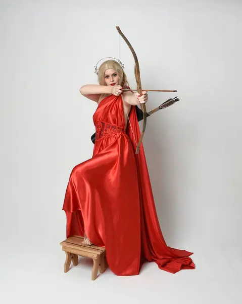 stock image Full length portrait of beautiful blonde model dressed as ancient mythological fantasy goddess in flowing red silk toga gown, crown. Graceful elegant pose  holding  archery bow and arrow weapons, isolated on white studio background.