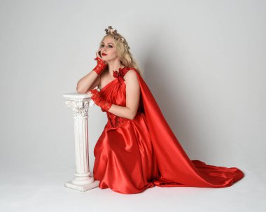 Eski mitolojik fantezi tanrıçası kılığında kırmızı ipek toga elbisesi ve taç giymiş güzel sarışın modelin tam boy portresi. Beyaz stüdyo arka planında izole edilmiş, Yunan sütunuyla diz çökmüş zarif bir poz..