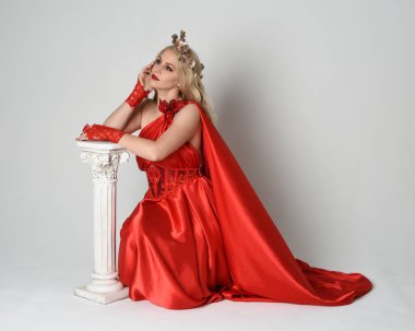 Eski mitolojik fantezi tanrıçası kılığında kırmızı ipek toga elbisesi ve taç giymiş güzel sarışın modelin tam boy portresi. Beyaz stüdyo arka planında izole edilmiş, Yunan sütunuyla diz çökmüş zarif bir poz..