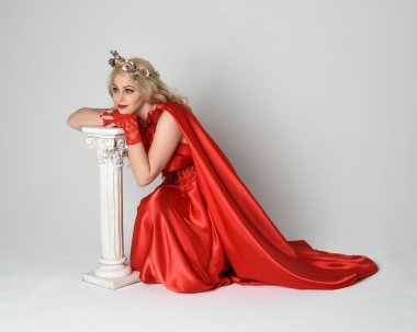 Eski mitolojik fantezi tanrıçası kılığında kırmızı ipek toga elbisesi ve taç giymiş güzel sarışın modelin tam boy portresi. Beyaz stüdyo arka planında izole edilmiş, Yunan sütunuyla diz çökmüş zarif bir poz..