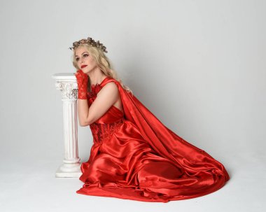 Eski mitolojik fantezi tanrıçası kılığında kırmızı ipek toga elbisesi ve taç giymiş güzel sarışın modelin tam boy portresi. Beyaz stüdyo arka planında izole edilmiş, Yunan sütunuyla diz çökmüş zarif bir poz..