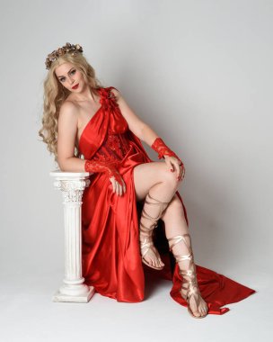 Eski mitolojik fantezi tanrıçası kılığında kırmızı ipek toga elbisesi ve taç giymiş güzel sarışın modelin tam boy portresi. Beyaz stüdyo arka planında izole edilmiş, Yunan sütunuyla diz çökmüş zarif bir poz..