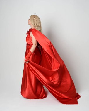 Eski mitolojik fantezi tanrıçası kılığında kırmızı ipek toga elbisesi ve taç giymiş güzel sarışın modelin tam boy portresi. Kameradan uzaklaşmak, beyaz stüdyo arka planında izole edilmiş..