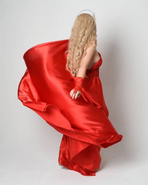 Eski mitolojik fantezi tanrıçası kılığında kırmızı ipek toga elbisesi ve taç giymiş güzel sarışın modelin tam boy portresi. Kameradan uzaklaşmak, beyaz stüdyo arka planında izole edilmiş..