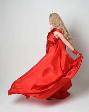 Eski mitolojik fantezi tanrıçası kılığında kırmızı ipek toga elbisesi ve taç giymiş güzel sarışın modelin tam boy portresi. Kameradan uzaklaşmak, beyaz stüdyo arka planında izole edilmiş..