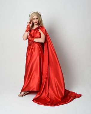 Eski mitolojik fantezi tanrıçası kılığında kırmızı ipek toga elbisesi ve taç giymiş güzel sarışın modelin tam boy portresi. Akan kumaşlı zarif dans pozu, beyaz stüdyo arka planında izole edilmiş..