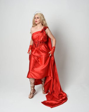 Eski mitolojik fantezi tanrıçası kılığında kırmızı ipek toga elbisesi ve taç giymiş güzel sarışın modelin tam boy portresi. Akan kumaşlı zarif dans pozu, beyaz stüdyo arka planında izole edilmiş..