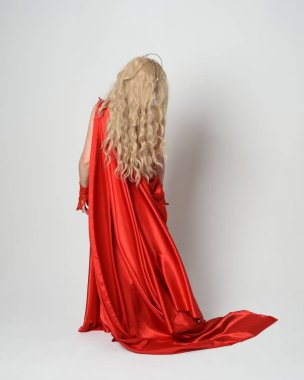 Eski mitolojik fantezi tanrıçası kılığında kırmızı ipek toga elbisesi ve taç giymiş güzel sarışın modelin tam boy portresi. Kameradan uzaklaşmak, beyaz stüdyo arka planında izole edilmiş..