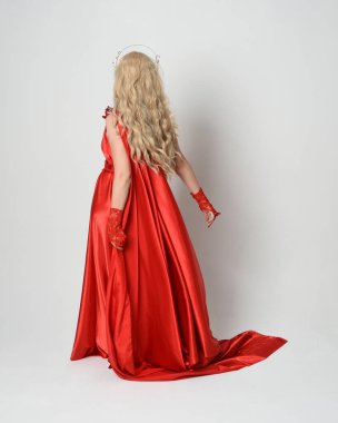 Eski mitolojik fantezi tanrıçası kılığında kırmızı ipek toga elbisesi ve taç giymiş güzel sarışın modelin tam boy portresi. Kameradan uzaklaşmak, beyaz stüdyo arka planında izole edilmiş..