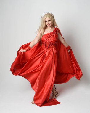 Eski mitolojik fantezi tanrıçası kılığında kırmızı ipek toga elbisesi ve taç giymiş güzel sarışın modelin tam boy portresi. Akan kumaşlı zarif dans pozu, beyaz stüdyo arka planında izole edilmiş..