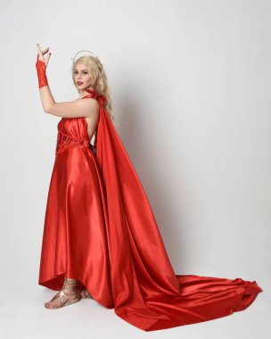 Eski mitolojik fantezi tanrıçası kılığında kırmızı ipek toga elbisesi ve taç giymiş güzel sarışın modelin tam boy portresi. Akan kumaşlı zarif dans pozu, beyaz stüdyo arka planında izole edilmiş..