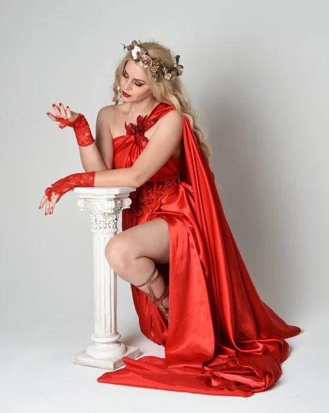 stock image Full length portrait of beautiful blonde model dressed as ancient mythological fantasy goddess in flowing red silk toga gown, crown. Graceful elegant pose kneeling with greek column, isolated on white studio background.