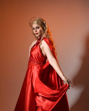 Akan kırmızı ipek toga elbisesi ve taç giyen, antik mitolojik fantezi tanrıçası Afrodit gibi giyinmiş güzel sarışın mankenin yakın portresi. Altın stüdyo arka planında izole edilmiş..