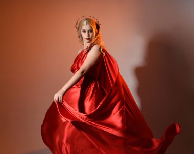 Akan kırmızı ipek toga elbisesi ve taç giyen, antik mitolojik fantezi tanrıçası Afrodit gibi giyinmiş güzel sarışın mankenin yakın portresi. Altın stüdyo arka planında izole edilmiş..