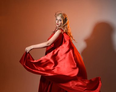 Akan kırmızı ipek toga elbisesi ve taç giyen, antik mitolojik fantezi tanrıçası Afrodit gibi giyinmiş güzel sarışın mankenin yakın portresi. Altın stüdyo arka planında izole edilmiş..