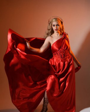 Akan kırmızı ipek toga elbisesi ve taç giyen, antik mitolojik fantezi tanrıçası Afrodit gibi giyinmiş güzel sarışın mankenin yakın portresi. Altın stüdyo arka planında izole edilmiş..
