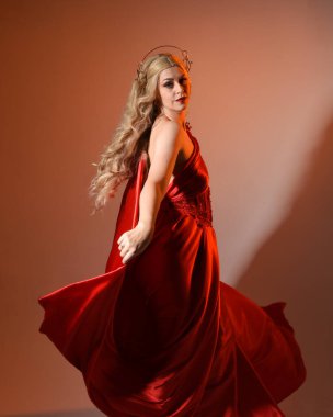 Akan kırmızı ipek toga elbisesi ve taç giyen, antik mitolojik fantezi tanrıçası Afrodit gibi giyinmiş güzel sarışın mankenin yakın portresi. Altın stüdyo arka planında izole edilmiş..