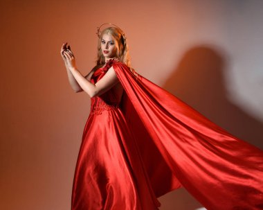 Akan kırmızı ipek toga elbisesi ve taç giyen, antik mitolojik fantezi tanrıçası Afrodit gibi giyinmiş güzel sarışın mankenin yakın portresi. Altın stüdyo arka planında izole edilmiş..