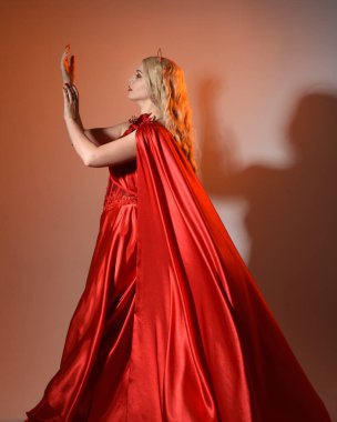 Akan kırmızı ipek toga elbisesi ve taç giyen, antik mitolojik fantezi tanrıçası Afrodit gibi giyinmiş güzel sarışın mankenin yakın portresi. Altın stüdyo arka planında izole edilmiş..