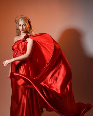 Akan kırmızı ipek toga elbisesi ve taç giyen, antik mitolojik fantezi tanrıçası Afrodit gibi giyinmiş güzel sarışın mankenin yakın portresi. Altın stüdyo arka planında izole edilmiş..