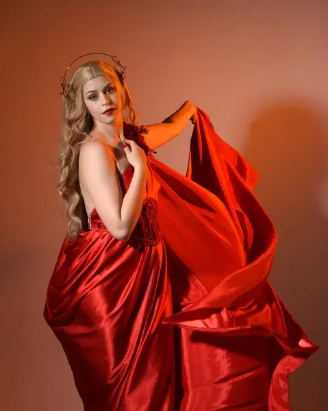Akan kırmızı ipek toga elbisesi ve taç giyen, antik mitolojik fantezi tanrıçası Afrodit gibi giyinmiş güzel sarışın mankenin yakın portresi. Altın stüdyo arka planında izole edilmiş..