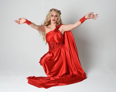 Eski mitolojik fantezi tanrıçası kılığında kırmızı ipek toga elbisesi ve taç giymiş güzel sarışın modelin tam boy portresi. Diz çök ve yere otur. Beyaz stüdyo arka planında izole.