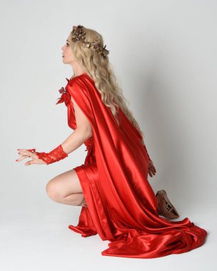 Eski mitolojik fantezi tanrıçası kılığında kırmızı ipek toga elbisesi ve taç giymiş güzel sarışın modelin tam boy portresi. Diz çök ve yere otur. Beyaz stüdyo arka planında izole.