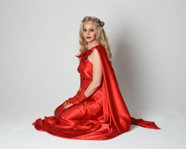 Eski mitolojik fantezi tanrıçası kılığında kırmızı ipek toga elbisesi ve taç giymiş güzel sarışın modelin tam boy portresi. Diz çök ve yere otur. Beyaz stüdyo arka planında izole.