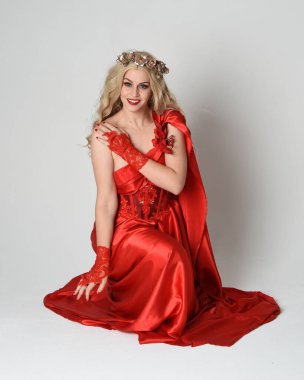 Eski mitolojik fantezi tanrıçası kılığında kırmızı ipek toga elbisesi ve taç giymiş güzel sarışın modelin tam boy portresi. Diz çök ve yere otur. Beyaz stüdyo arka planında izole.