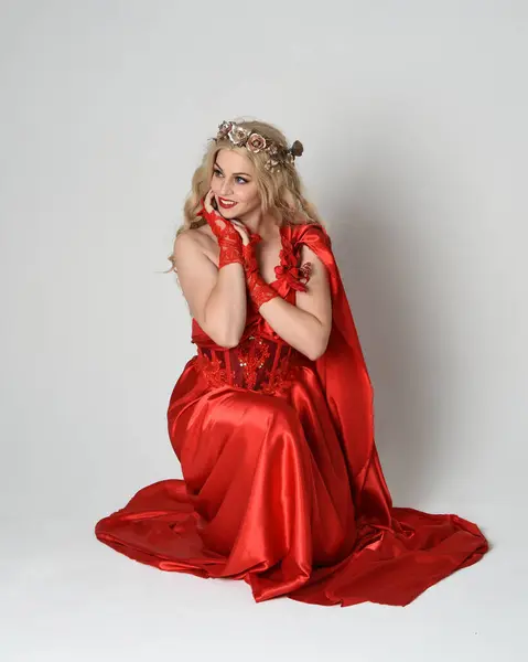 stock image Full length portrait of beautiful blonde model dressed as ancient mythological fantasy goddess in flowing red silk toga gown, crown. Kneeling pose sitting on floor. isolated on white studio background.