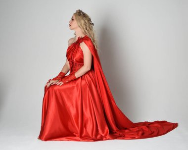 Eski mitolojik fantezi tanrıçası kılığında kırmızı ipek toga elbisesi ve taç giymiş güzel sarışın modelin tam boy portresi. Sandalyeye oturmuş poz veriyor. Beyaz stüdyo arka planında izole.