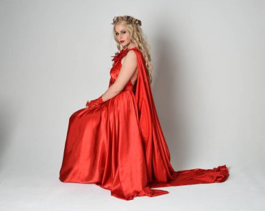 Eski mitolojik fantezi tanrıçası kılığında kırmızı ipek toga elbisesi ve taç giymiş güzel sarışın modelin tam boy portresi. Sandalyeye oturmuş poz veriyor. Beyaz stüdyo arka planında izole.