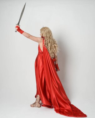 Eski mitolojik fantezi tanrıçası kılığında kırmızı ipek toga elbisesi ve taç giymiş güzel sarışın modelin tam boy portresi. Arkadan bakmak, elinde kılıçla yürüyüp gitmek, beyaz stüdyo arka planında izole edilmiş..