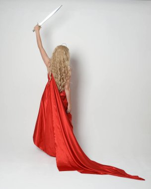 Eski mitolojik fantezi tanrıçası kılığında kırmızı ipek toga elbisesi ve taç giymiş güzel sarışın modelin tam boy portresi. Arkadan bakmak, elinde kılıçla yürüyüp gitmek, beyaz stüdyo arka planında izole edilmiş..