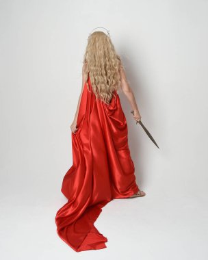 Eski mitolojik fantezi tanrıçası kılığında kırmızı ipek toga elbisesi ve taç giymiş güzel sarışın modelin tam boy portresi. Arkadan bakmak, elinde kılıçla yürüyüp gitmek, beyaz stüdyo arka planında izole edilmiş..