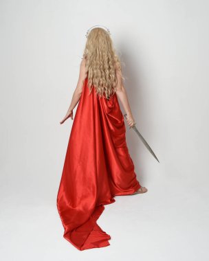 Eski mitolojik fantezi tanrıçası kılığında kırmızı ipek toga elbisesi ve taç giymiş güzel sarışın modelin tam boy portresi. Arkadan bakmak, elinde kılıçla yürüyüp gitmek, beyaz stüdyo arka planında izole edilmiş..