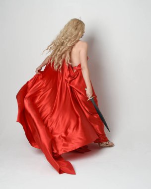 Eski mitolojik fantezi tanrıçası kılığında kırmızı ipek toga elbisesi ve taç giymiş güzel sarışın modelin tam boy portresi. Arkadan bakmak, elinde kılıçla yürüyüp gitmek, beyaz stüdyo arka planında izole edilmiş..