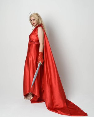 Eski mitolojik fantezi tanrıçası kılığında kırmızı ipek toga elbisesi ve taç giymiş güzel sarışın modelin tam boy portresi. Yürüyen bir poz, kılıç tutan bir silah, izole edilmiş bir stüdyo.