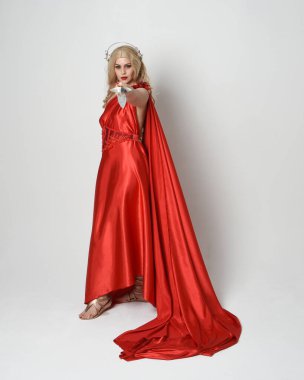 Eski mitolojik fantezi tanrıçası kılığında kırmızı ipek toga elbisesi ve taç giymiş güzel sarışın modelin tam boy portresi. Yürüyen bir poz, kılıç tutan bir silah, izole edilmiş bir stüdyo.