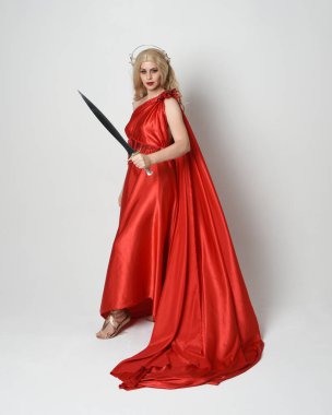 Eski mitolojik fantezi tanrıçası kılığında kırmızı ipek toga elbisesi ve taç giymiş güzel sarışın modelin tam boy portresi. Yürüyen bir poz, kılıç tutan bir silah, izole edilmiş bir stüdyo.