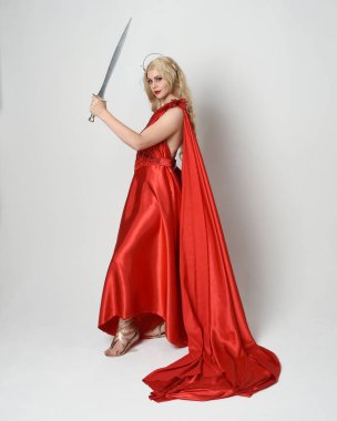 Eski mitolojik fantezi tanrıçası kılığında kırmızı ipek toga elbisesi ve taç giymiş güzel sarışın modelin tam boy portresi. Yürüyen bir poz, kılıç tutan bir silah, izole edilmiş bir stüdyo.