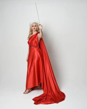 Eski mitolojik fantezi tanrıçası kılığında kırmızı ipek toga elbisesi ve taç giymiş güzel sarışın modelin tam boy portresi. Yürüyen bir poz, kılıç tutan bir silah, izole edilmiş bir stüdyo.