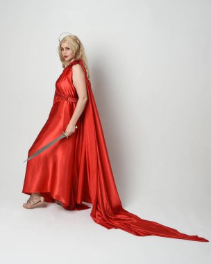 Eski mitolojik fantezi tanrıçası kılığında kırmızı ipek toga elbisesi ve taç giymiş güzel sarışın modelin tam boy portresi. Yürüyen bir poz, kılıç tutan bir silah, izole edilmiş bir stüdyo.